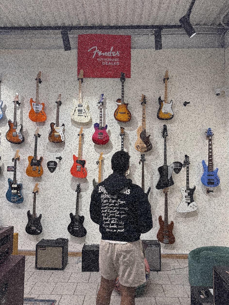 Sahil from behind, facing a shop wall hung with electric guitars under a Fender sign